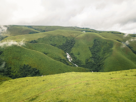 Aerial view of lush undulating emerald hills embracing a silver ribbon waterfall cascading through verdant valleys under a soft cloudy sky Obudu Cross River Nigeria.