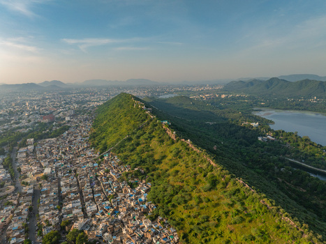 Aerial view of the historic city of Udaipur featuring a prominent green hill ridge separating dense urban rooftops from the serene lake under a clear blue sky. Udaipur Rajasthan India.