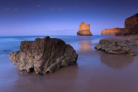 View of limestone stacks of the Twelve Apostles Marine National Park at twilight with large rocks on a sandy beach and calm ocean water in Australia. View of limestone stacks of the Twelve Apostles Marine National Park at twilight with large rocks on a sandy beach and calm ocean water in Australia.