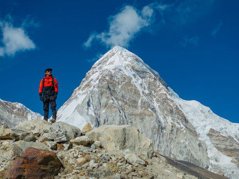 Aerial view of a hiker in an orange jacket standing on a rocky slope with a snow capped mountain peak in the background under a clear blue sky in Nepal.