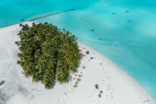 Aerial view of Aitutaki with lush palm trees on a white sand beach surrounded by turquoise lagoon waters and kitesurfers in Cook Islands. Aerial view of Aitutaki with lush palm trees on a white sand beach surrounded by turquoise lagoon waters and kitesurfers in Cook Islands.