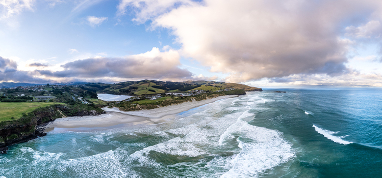 Aerial view of the rugged coastline near Dunedin featuring sandy beaches rolling green hills crashing ocean waves and dramatic cliffs in Dunedin Otago Region New Zealand. Aerial view of the rugged coastline near Dunedin featuring sandy beaches rolling green hills crashing ocean waves and dramatic cliffs in Dunedin Otago Region New Zealand.