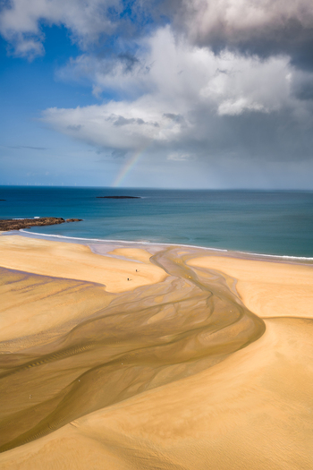 Aerial view of a wide sandy beach with a river estuary flowing into the blue sea under a dramatic sky with a rainbow Plurien Brittany France. Aerial view of a wide sandy beach with a river estuary flowing into the blue sea under a dramatic sky with a rainbow Plurien Brittany France.