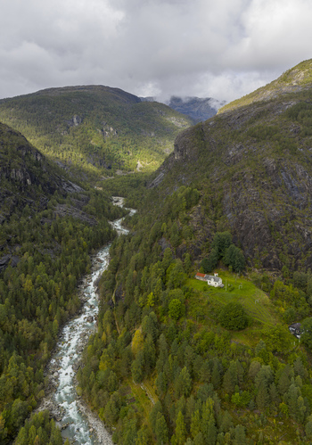 Aerial view of a winding river flowing through a lush valley with a solitary white house on a green hillside and steep rocky mountains under a cloudy sky Fjaera Vestland Norway. Aerial view of a winding river flowing through a lush valley with a solitary white house on a green hillside and steep rocky mountains under a cloudy sky Fjaera Vestland Norway.