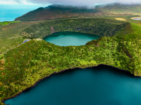 Aerial view of a vibrant blue lake nestled within a lush green volcanic crater contrasting with the deep blue ocean in the distance Flores Azores Portugal.