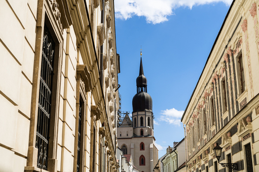 View of the historic St. Nicholas Basilica tower framed by ornate classical facades along a narrow cobblestone street under a bright blue sky Trnava Trnava Region Slovakia. View of the historic St. Nicholas Basilica tower framed by ornate classical facades along a narrow cobblestone street under a bright blue sky Trnava Trnava Region Slovakia.