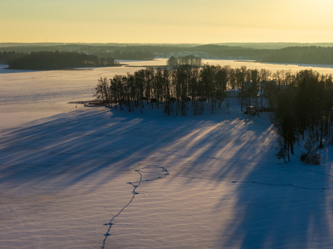 Aerial view of a frozen lake and a snow covered island with bare trees casting long shadows during golden hour Lithuania. Aerial view of a frozen lake and a snow covered island with bare trees casting long shadows during golden hour Lithuania.