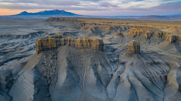 Aerial view of the rugged badlands landscape with towering sandstone formations deep erosion gullies and distant mountains under a soft sunset sky in Hanksville Utah United States.