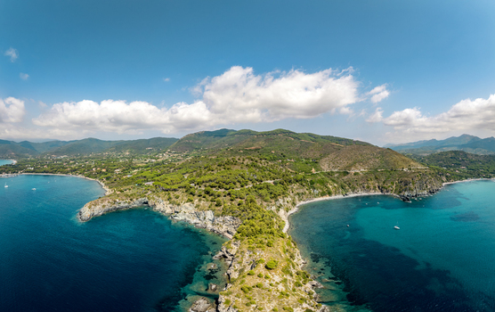 Aerial view of Lacona Beach with turquoise waters rocky cliffs and lush green hills under a bright blue sky in Lacona Tuscany Italy.