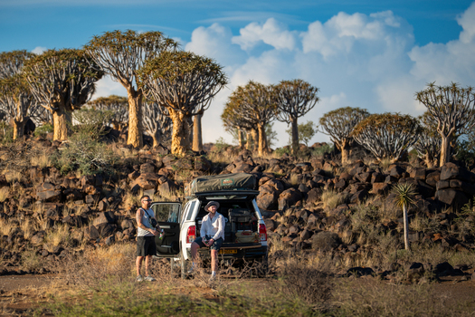 Aussenkehr Namibia 06 March 2026 View of two figures pause by a white vehicle with a rooftop tent amidst a landscape of quiver trees and dark rugged rocks under a sky streaked with wispy clouds. Aussenkehr Namibia 06 March 2026 View of two figures pause by a white vehicle with a rooftop tent amidst a landscape of quiver trees and dark rugged rocks under a sky streaked with wispy clouds.