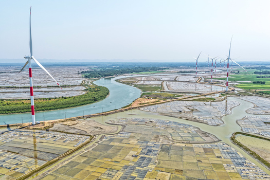 Aerial view of towering wind turbines with red and white stripes stand amidst a patchwork of flooded fields and a winding river Cox s Bazar Chittagong Division Bangladesh. Aerial view of towering wind turbines with red and white stripes stand amidst a patchwork of flooded fields and a winding river Cox s Bazar Chittagong Division Bangladesh.