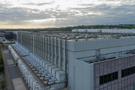 Aerial view of a large data center facility featuring rows of industrial cooling units on the roof under a cloudy sunset sky in the United Kingdom.