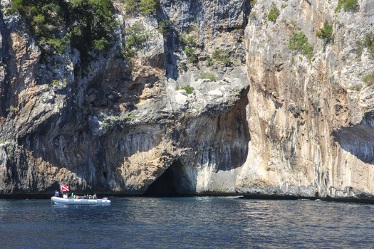 View of a small boat with passengers near the towering limestone cliffs and sea caves of the Gulf of Orosei Italy. View of a small boat with passengers near the towering limestone cliffs and sea caves of the Gulf of Orosei Italy.