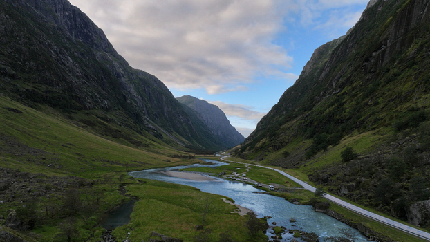 Aerial view of a winding river flowing through a lush green valley flanked by towering mountains under a cloudy sky with a road and parked vehicles in Nordland Norway. Aerial view of a winding river flowing through a lush green valley flanked by towering mountains under a cloudy sky with a road and parked vehicles in Nordland Norway.