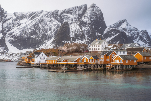 View of vibrant yellow and white wooden houses perched on stilts above the tranquil waters against a backdrop of snow dusted mountains Hamn&oslash;y Nordland Norway.