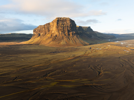 Aerial view of Lomagnupur mountain rising above the vast volcanic plains with golden sunlight illuminating the rugged cliffs and dark sandy landscape in Skaftarhreppur Iceland.