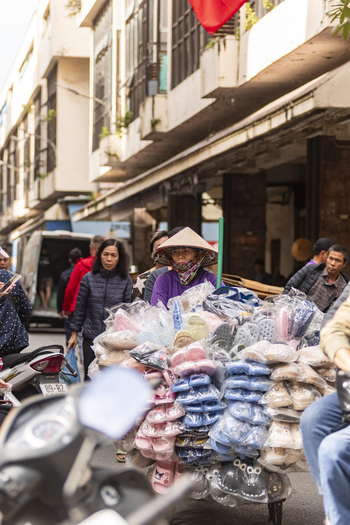 Hanoi Ha Noi Vietnam 11 November 2025 View of a street vendor wearing a conical hat pushing a cart filled with plastic footwear through a busy street.