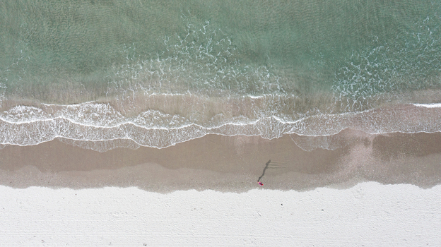 Aerial view of Omaha Beach with white sand gentle waves washing the shore and a person walking along the coastline in Omaha Auckland New Zealand.