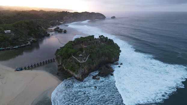 Aerial view of Pantai Drini with its rocky island wooden bridge sandy beach and crashing waves during sunset in Kecamatan Tanjungsari Daerah Istimewa Yogya Indonesia. Aerial view of Pantai Drini with its rocky island wooden bridge sandy beach and crashing waves during sunset in Kecamatan Tanjungsari Daerah Istimewa Yogya Indonesia.