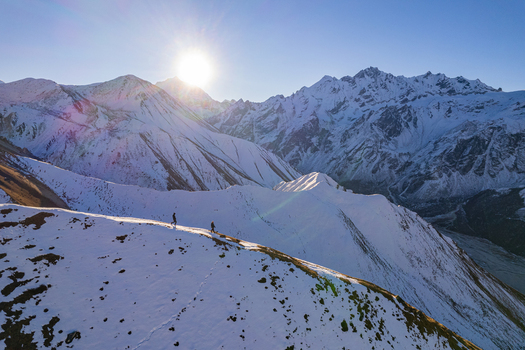 Aerial view of two hikers walking along a snow covered mountain ridge in the Himalayas under a bright sun with vast snow capped peaks in the background in Nepal.