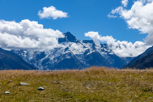 View of snow capped mountains piercing the bright blue sky partially veiled by puffy white clouds above a field of golden grass Mount Cook Canterbury Region New Zealand. View of snow capped mountains piercing the bright blue sky partially veiled by puffy white clouds above a field of golden grass Mount Cook Canterbury Region New Zealand.