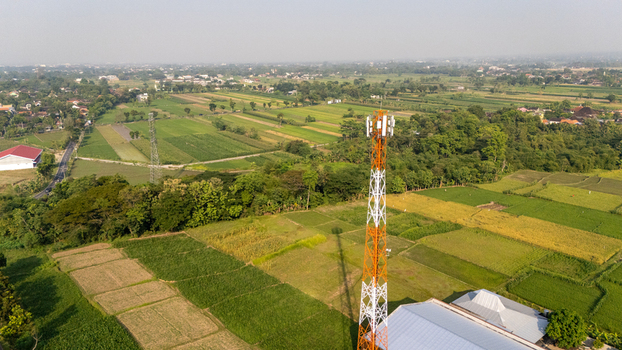 Aerial view of a tall telecommunications tower standing in a lush green agricultural landscape with rice fields and scattered trees in Indonesia.