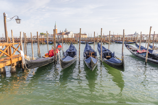 View of traditional gondolas moored to wooden posts in the calm waters of the lagoon with historic buildings in the background in Venice Veneto Italy. View of traditional gondolas moored to wooden posts in the calm waters of the lagoon with historic buildings in the background in Venice Veneto Italy.