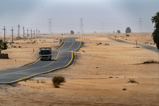 View of a white truck driving on a winding asphalt road through the desert dunes with power lines in the distance Dubai Dubai United Arab Emirates. View of a white truck driving on a winding asphalt road through the desert dunes with power lines in the distance Dubai Dubai United Arab Emirates.