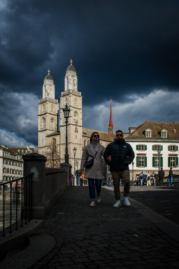 Zurich Switzerland 02 April 2023 View of a couple strolling across a bridge with the imposing Grossmünster church silhouetted against a dark stormy sky. Zurich Switzerland 02 April 2023 View of a couple strolling across a bridge with the imposing Grossmünster church silhouetted against a dark stormy sky.