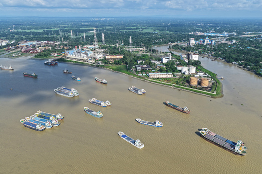 Aerial view of Karnaphuli River with numerous cargo ships and barges navigating the waterway near industrial facilities and lush greenery in Chattogram Chittagong Division Bangladesh.