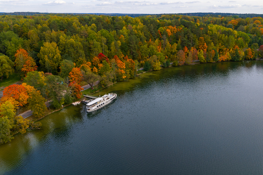 Aerial view of a white passenger boat docked at a pier on the shore of a lake surrounded by a dense forest with vibrant autumn foliage in Trakai Vilnius County Lithuania. Aerial view of a white passenger boat docked at a pier on the shore of a lake surrounded by a dense forest with vibrant autumn foliage in Trakai Vilnius County Lithuania.