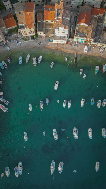 Aerial view of small boats moored in the turquoise harbor waters next to historic stone houses and a pebble beach in Komiza Split Dalmatia County Croatia.