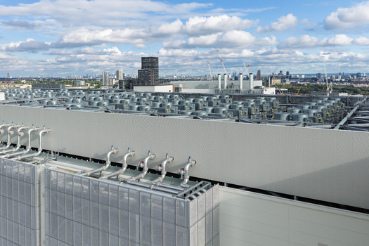 Aerial view of a data center rooftop featuring rows of cooling units and industrial piping with a city skyline in the background United Kingdom. Aerial view of a data center rooftop featuring rows of cooling units and industrial piping with a city skyline in the background United Kingdom.