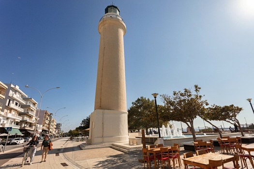 View of the Alexandroupoli Lighthouse on the waterfront with outdoor cafe seating trees and people walking along the sidewalk under a clear blue sky in Alexandroupoli Greece. View of the Alexandroupoli Lighthouse on the waterfront with outdoor cafe seating trees and people walking along the sidewalk under a clear blue sky in Alexandroupoli Greece.