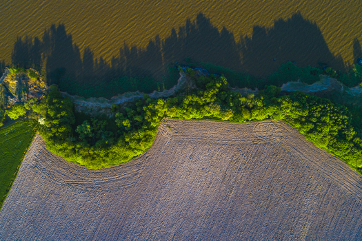Aerial view of a cultivated field bordering a lake with lush green trees and long shadows at sunset in Vanersnas Vastra Gotalands lan Sweden. Aerial view of a cultivated field bordering a lake with lush green trees and long shadows at sunset in Vanersnas Vastra Gotalands lan Sweden.