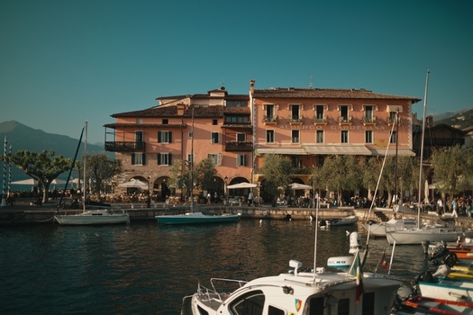 View of Lake Garda waterfront featuring the historic Albergo Gardesana building and boats moored in the harbor against a mountain backdrop in Italy. View of Lake Garda waterfront featuring the historic Albergo Gardesana building and boats moored in the harbor against a mountain backdrop in Italy.
