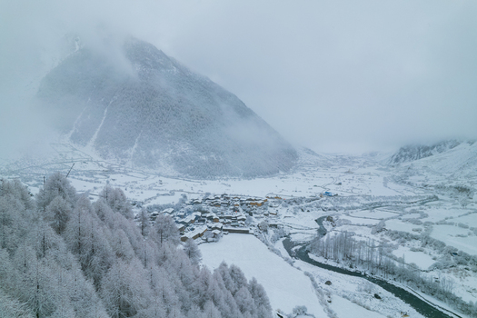 Aerial view of a snow covered village nestled in a mountain valley with a winding river and frost covered trees under a cloudy sky in Nepal.