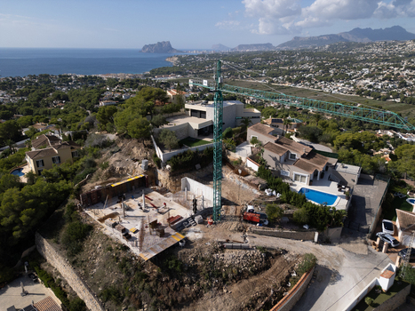 Aerial view of a construction site with a tall green crane concrete foundations and surrounding villas with swimming pools overlooking the coastline in Moraira Valencian Community Spain.