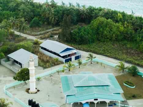 Aerial view of mosque with tall minaret and nearby buildings nestled amid lush greenery where the land meets the turquoise sea Vashafaru Island Maldives. Aerial view of mosque with tall minaret and nearby buildings nestled amid lush greenery where the land meets the turquoise sea Vashafaru Island Maldives.