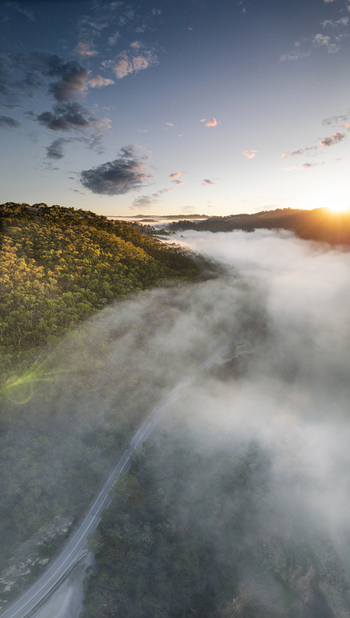 Aerial view of Kangaroo Creek Reservoir area with a winding road emerging from thick morning fog and golden sunrise light over forested hills Adelaide Hills Council South Australia Australia. Aerial view of Kangaroo Creek Reservoir area with a winding road emerging from thick morning fog and golden sunrise light over forested hills Adelaide Hills Council South Australia Australia.
