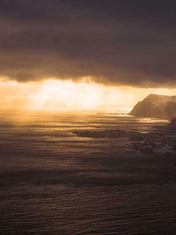 Aerial view of shimmering waters reflecting the golden light under a dramatic sky meeting the rugged coastline Reinebringen Nordland Norway.