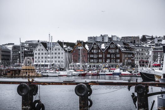 View of the harbor with traditional wooden buildings and boats docked along the waterfront under an overcast sky in Tromso Troms Norway. View of the harbor with traditional wooden buildings and boats docked along the waterfront under an overcast sky in Tromso Troms Norway.