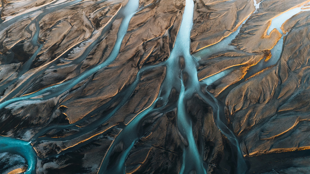 Aerial view of intricate braided glacial river patterns flowing through dark volcanic sand with golden sunlight highlights Skaftarhreppur Iceland.