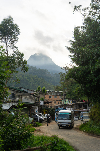 View of Adam s Peak rising above the village buildings and a road with local traffic under a cloudy sky in Nallathanniya Central Province Sri Lanka. View of Adam s Peak rising above the village buildings and a road with local traffic under a cloudy sky in Nallathanniya Central Province Sri Lanka.