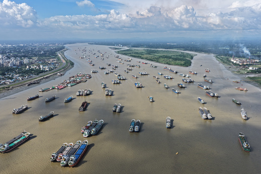 Aerial view of Karnaphuli River filled with numerous cargo ships and barges with urban skyline and lush green banks in Chattogram Chattogram Division Bangladesh.