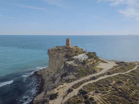 Aerial view of Torre del Xarco standing on a rugged cliff overlooking the turquoise Mediterranean Sea under a clear blue sky in La Vila Joiosa Comunidad Valenciana Spain.