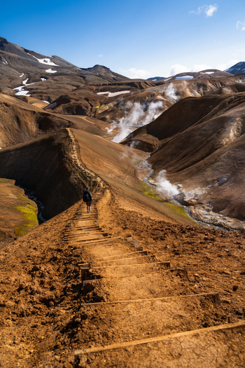 View of a solitary figure traversing a rugged ochre toned trail amidst steaming geothermal vents and snow capped peaks under a clear sky Hrunamannahreppur Iceland.
