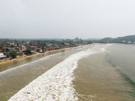 Aerial view of Dewata Beach Road featuring a long sandy coastline with breaking waves and coastal buildings under a hazy sky in Galle Southern Province Sri Lanka.