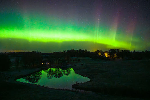 Aerial view of the aurora borealis illuminating the night sky with green and purple lights reflecting over a calm pond surrounded by dark trees and a small building in Latvia. Aerial view of the aurora borealis illuminating the night sky with green and purple lights reflecting over a calm pond surrounded by dark trees and a small building in Latvia.