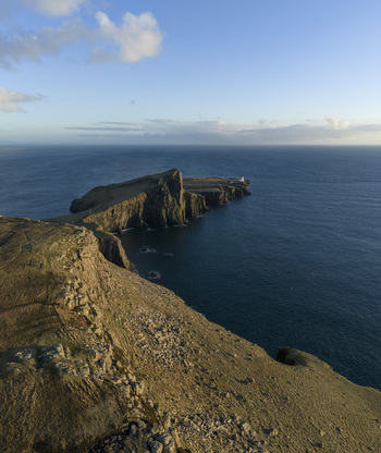 Aerial view of Neist Point Lighthouse on a rocky headland with steep cliffs overlooking the deep blue Atlantic Ocean under a blue sky in Isle of Skye Scotland United Kingdom. Aerial view of Neist Point Lighthouse on a rocky headland with steep cliffs overlooking the deep blue Atlantic Ocean under a blue sky in Isle of Skye Scotland United Kingdom.